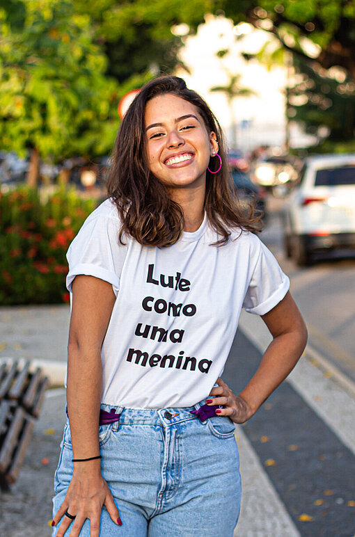 Luiza war schon immer an dem Thema Feminismus interessiert. Luisa steht mit einem breiten Lächeln und einer in die Hüfte gestemmten Hand auf einem Gehweg.