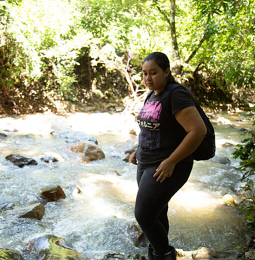 Eine junge Frau steht vor einem breiten Bach, in dem das Wasser stark fließt