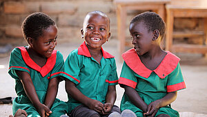 Drei lachende Kinder aus Malawi in grünen Schuluniformen mit roten Kragen sitzen nebeneinander und schauen sich fröhlich an.