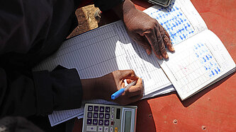 BR dokumentiert die Einzahlungen und die Kredite. Neben Heften und Listen liegt ein Taschenrechner auf dem Tisch.