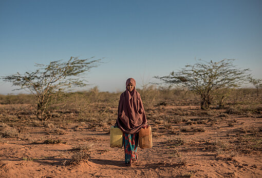 Eine Elfjährige aus Somalia läuft durch trockene Landschaft mit zwei Wasserkanistern in der Hand