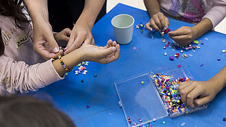 Kinder basteln bunte Armbänder aus Perlen; eine erwachsene Person hilft dabei, ein Armband am Handgelenk eines Kindes zu befestigen.