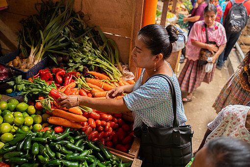 Eine Frau steht an einem Marktstand voller Gemüse und hält eine Paprika in der Hand.