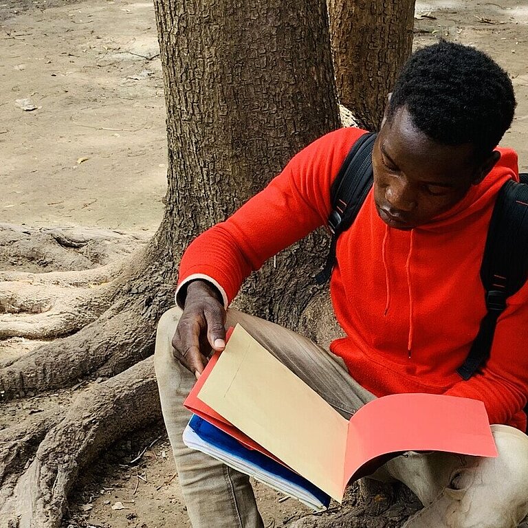 Ein Junge sitzt an einen Baum gelehnt auf dem Boden und liest in einem Schulheft
