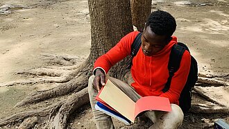 Ein Junge sitzt an einen Baum gelehnt auf dem Boden und liest in einem Schulheft