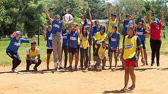 Ein Mädchen steht auf einem Sandplatz und schießt einen Ball, am Spielfeldrand stehen Jungen und Mädchen und feuern die Spielerin an