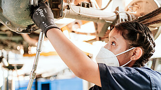 Eine junge Frau, die eine FFP2-Maske trägt, steht unter einem Autoteil und schraubt etwas daran