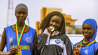 Drei junge Senegalesinnen stehen auf einem Podest, jede hat eine Medaille umhängen. Das Mädchen in der Mitte beißt auf ihre Medaille.