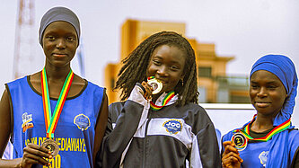 Drei junge Senegalesinnen stehen auf einem Podest, jede hat eine Medaille umhängen. Das Mädchen in der Mitte beißt auf ihre Medaille.