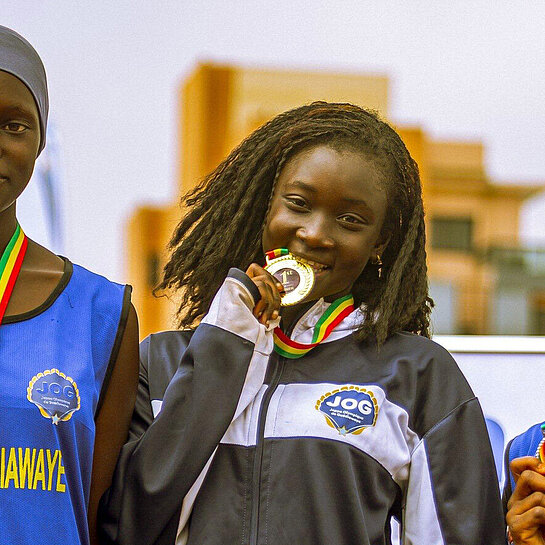 Drei junge Senegalesinnen stehen auf einem Podest, jede hat eine Medaille umhängen. Das Mädchen in der Mitte beißt auf ihre Medaille.