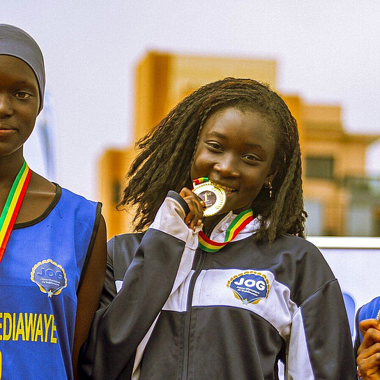 Drei junge Senegalesinnen stehen auf einem Podest, jede hat eine Medaille umhängen. Das Mädchen in der Mitte beißt auf ihre Medaille.