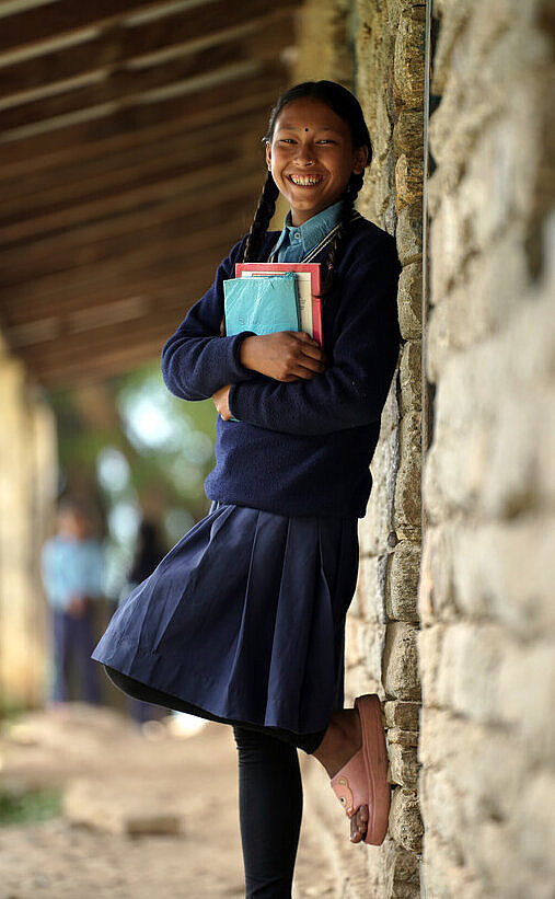 Die Schulen im Distrikt Kalikot sind heute ein positiverer Ort für Mädchen, dank gezielter Bildungsprogramme und Sensibilisierungskampagnen Eine Schülerin aus Nepal lächelt und trägt Schulmaterialien in ihrer Hand