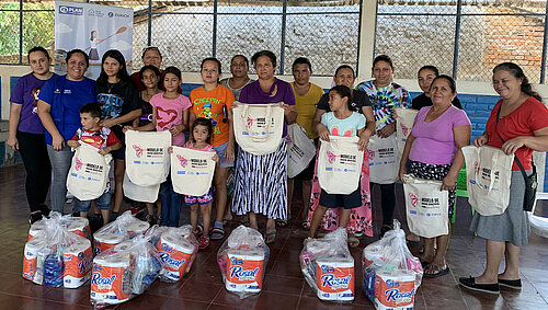 Eine Gruppe aus Frauen, Kindern und Jugendlichen steht in einer Reihe in einer offenen Halle, die Pakete mit Lebensmitteln und Hygieneartikeln erhalten hat. Die Teilnehmer sind in bunter Alltagskleidung gekleidet und halten Stofftaschen.