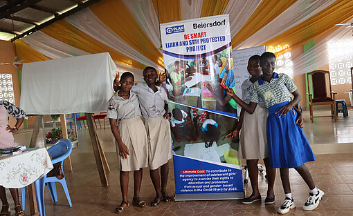 Mädchen bei einem SGBV-Workshop in Ghana. Eine Gruppe Mädchen steht vor dem Banner des Workshops, auf dem das Plan und das Beiersdorf-Logo zu sehen ist.