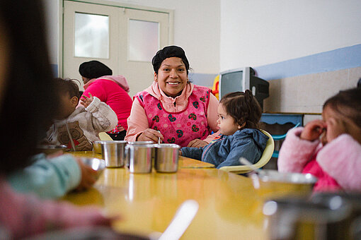 Eine Betreuerin in einem rosa Kittel sitzt mit Kleinkindern an einem Tisch und hilft ihnen beim Essen. Die Kinder schauen auf Teller, die vor ihnen stehen.