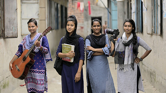 Eine Gruppe Mädchen steht in einer Gasse. In ihren Häden halten sie eine Kamera, eine Gitarre, und ein Buch.