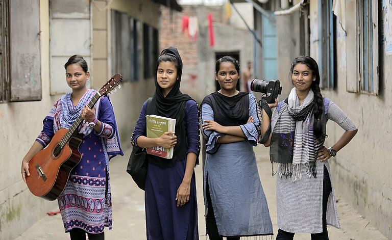 Eine Gruppe Mädchen steht in einer Gasse und schaut selbstbewusst in die Kamera. Sie tragen eine Gitarre, ein Buch und eine Kamera bei sich.