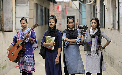 Auf dem Bild ist eine Gruppe Mädchen zu sehen. In den Händern halten sie eine Gitarre, ein Buch und eine Kamera. Sie schauen selbstbewusst und verschmitzt in die Kamera.