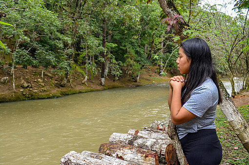 Anahi blickt auf den Fluss, der neben ihrem Zuhause verläuft Anahi steht mit verschränkten Händen am Flussufer und blickt auf das Wasser