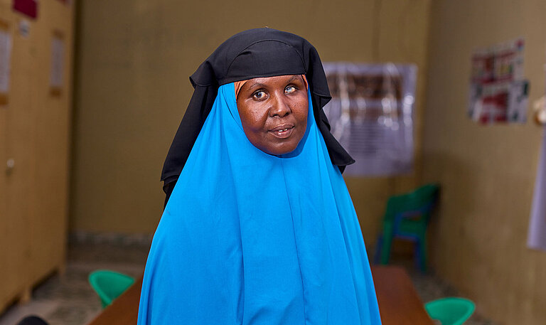 Eine Frau in leuchtend blauem Gewand und schwarzem Kopftuch schaut in die Kamera. Sie steht vor einem Konferenztisch.
