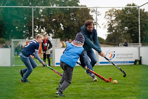 Zwei Kinder und ein Erwachsener spielen Feldhockey, im Hintergrund steht einweiteres Kind im Tor