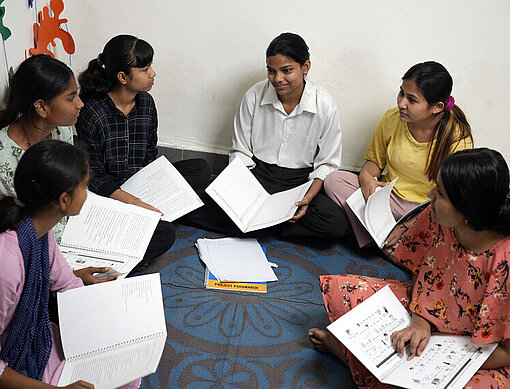 Anjali (Mitte) nimmt mit ihren Freundinnen an einem Workshop im Trainingszentrum von Plan International teil Eine Gruppe junger Frauen sitzt auf dem Boden im Kreis und tauscht sich aus. Sie haben alle Hefte und Stifte vor sich.