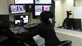 Eine Frau sitzt vor Monitoren und einem Schaltpult.