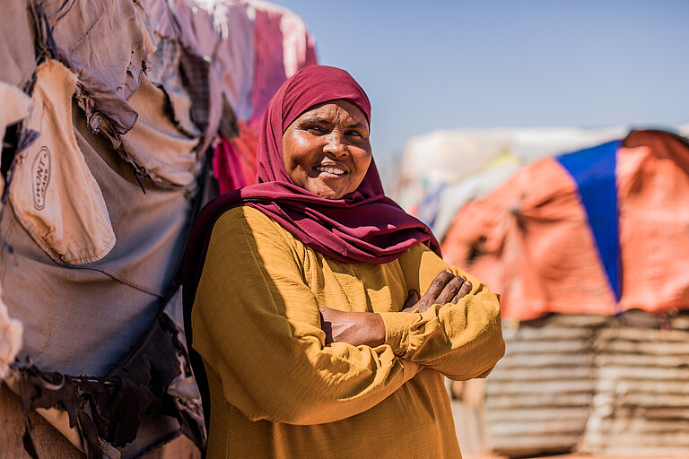 Saafi steht vor einer Hütte im Camp für Binnenvertriebene. Sie hat die Arme vor der Brust verschränkt, lächelt in die Kamera.