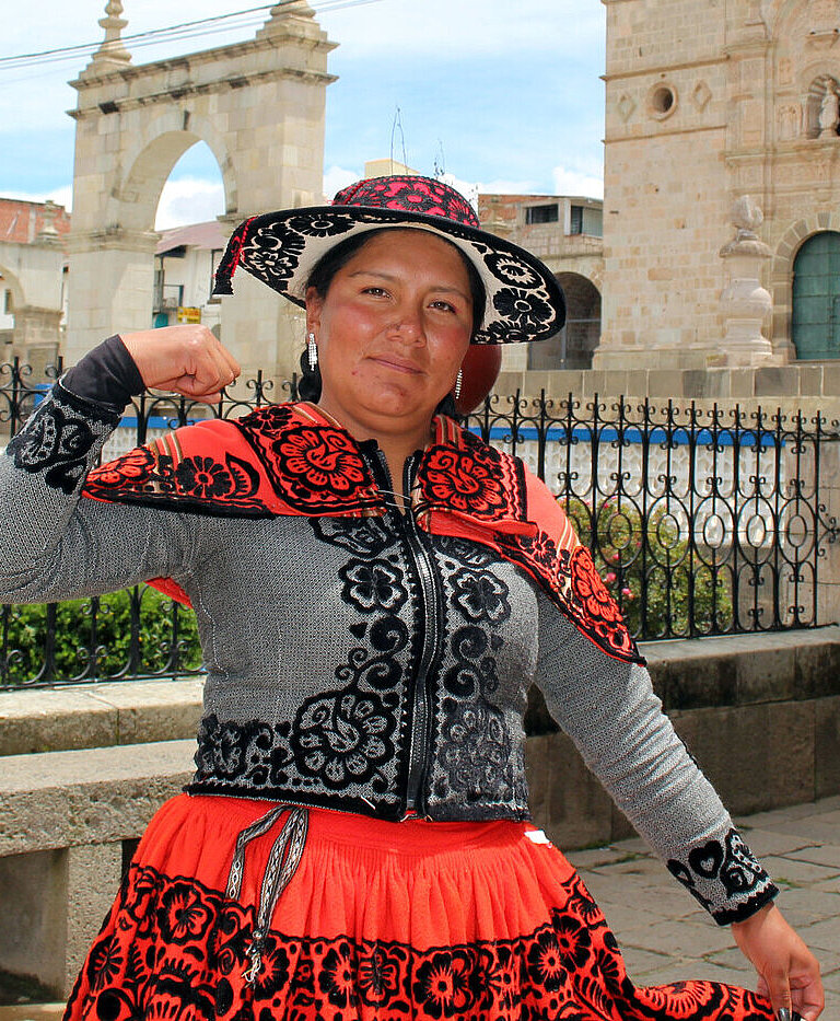 Jessica aus Chumbivilcas, Peru, steht vor einer historischen Kirche und zeigt stolz ihre Stärke, während sie traditionelle Kleidung trägt.