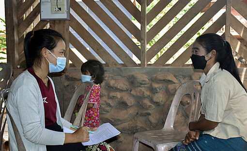 Zwei Frauen, die Mund-Nasen-Masken tragen, sitzen sich gegenüber, eine von ihnen hält einen Block und Stift in der Hand.