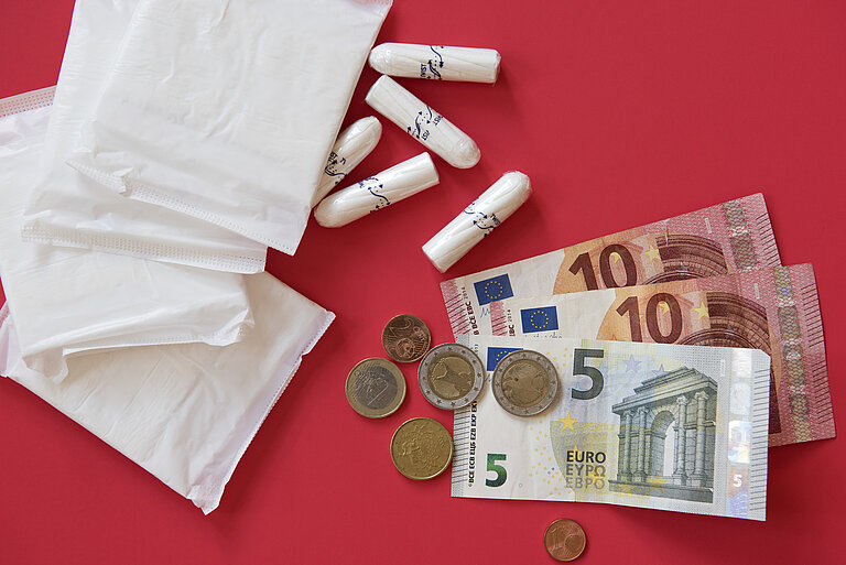 Auf einer Fläche liegen zwei Binden, ein paar Tampons, ein Kassenzettel und Geldscheine