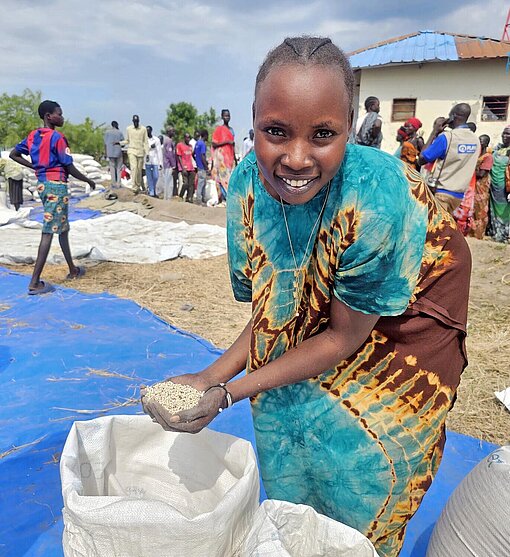 Eine junge Frau in buntem Kleid zeigt einen Sach mit Getreide