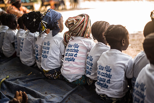 FGM/C Eine Gruppe junger Mädchen sitzt nebeneinander, die Rücken zur Kamera. Sie tragen alle das gleiche T-Shirt mit einem Druck auf dem Rücken in französischer Sprache.
