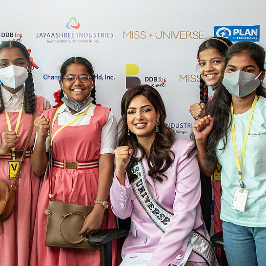 Eine Gruppe Mädchen steht neben einer Frau, die eine Miss Universe-Schärpe trägt und eine Faust für Empowerment ballt