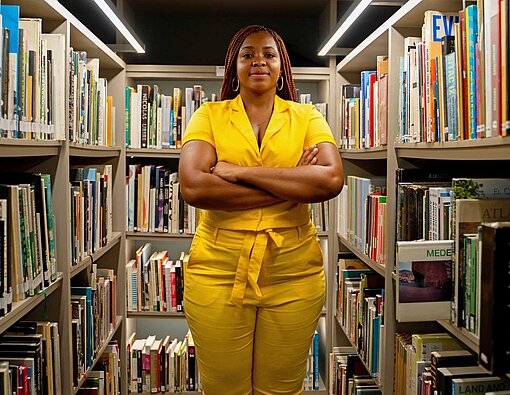 Eine dunkelhäutige Frau in leuchtend gelbem Kostüm steht zwischen zwei Bücherregalen in einer Bibliothek.