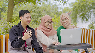 Ein junger Mann und zwei junge Frauen sitzen vor einem Laptop und schauen lächelnd darau