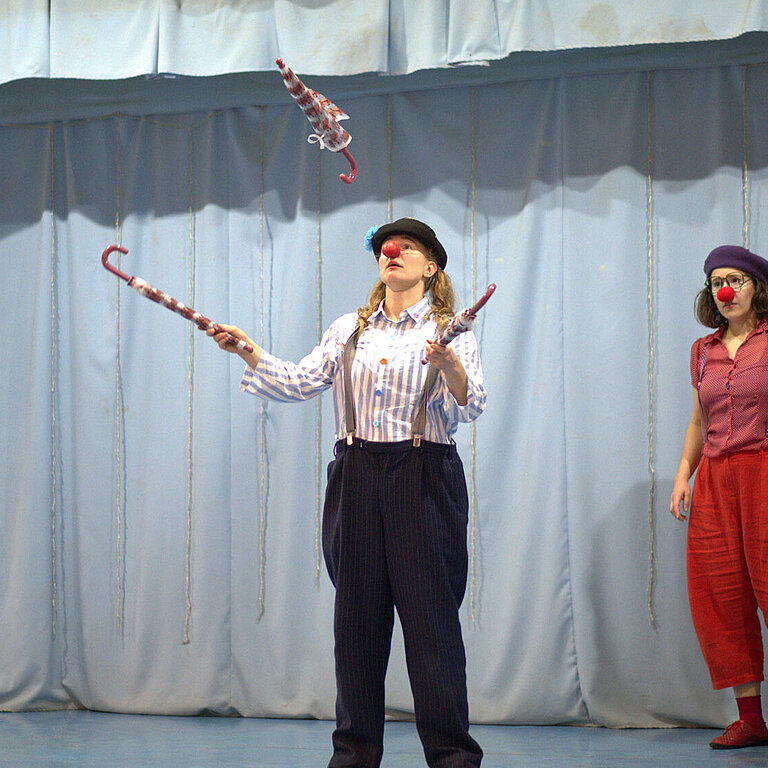 Drei Clowns stehen auf einer Bühne. Eine von ihnen jongliert mit drei Regenschirmen.
