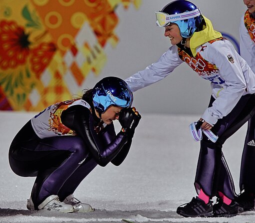 Eine Skispringerin ist in der Hocke und hat die Fäuste an die Stirn gelegt, eine andere Skispringerin steht vor ihr, lächelt und hat die rechte Hand auf die Schulter der anderen Skispringerin gelegt
