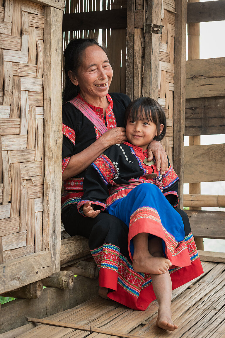 Eine Frau sitzt in einem Hauseingang und lacht, sie hat ein lächelndes Mädchen auf dem Schoß. Beide tragen traditionelle, schwarz und rot bestickte Kleider.
