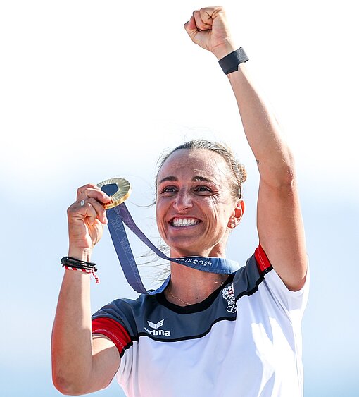 Als Profiseglerin gewann Lara Vadlau die Welt- und Europameisterschaften sowie bei den Olympischen Spielen 2024 die Goldmedaille Lara Vadlau hält mit der einen Hand ihre Olympia-Medaille neben ihren Kopf und streckt die andere Hand in einer Siegesgeste nach oben