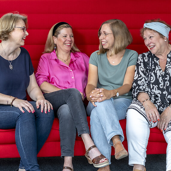 Vier Frauen sitzen auf einem roten Sofa und lachen.