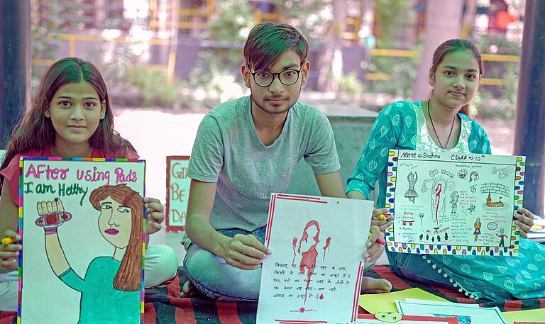 Kinder sitzen in Indien unter einer Zeltplane auf Decken und zeigen Plakate über Menstruationshygiene vor