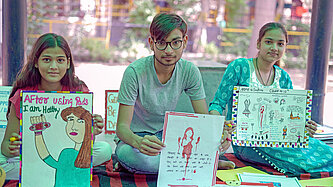 Kinder sitzen in Indien unter einer Zeltplane auf Decken und zeigen Plakate über Menstruationshygiene vor