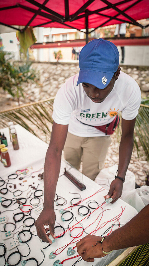 Ein junger Mann steht an einem Marktstand und zeigt einem Kunden ein handgefertigtes Kunstobjekt.