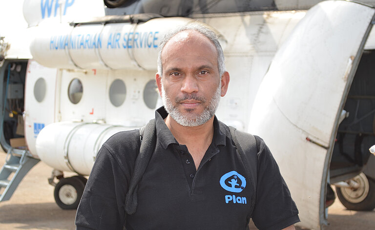 Dr. Unni Krishnan steht vor einem Flugzeug. Auf seinem T-Shirt ist das Plan International Logo