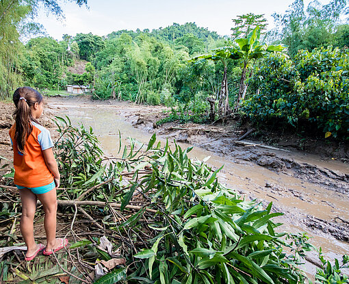 Ein Mädchen steht vor einer schlammigen Straße, die durch Überschwemmungen unbefahrbar geworden ist