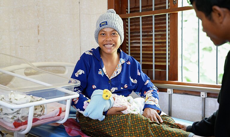 Eine frisch entbundene Mutter sitzt im Gesundheitszentrum, hält ihr Baby im Arm und lächelt glücklich in die Kamera.
