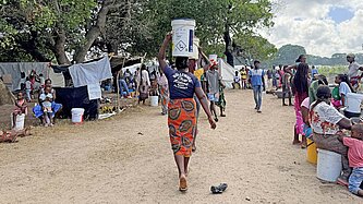 Eine Frau balanciert Güter auf dem Kopf und läuft durch ein Notcamp aus Zelten