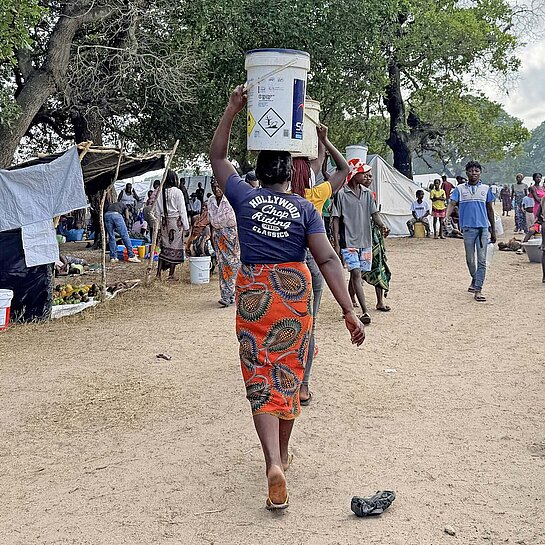 Eine Frau balanciert Güter auf dem Kopf und läuft durch ein Notcamp aus Zelten