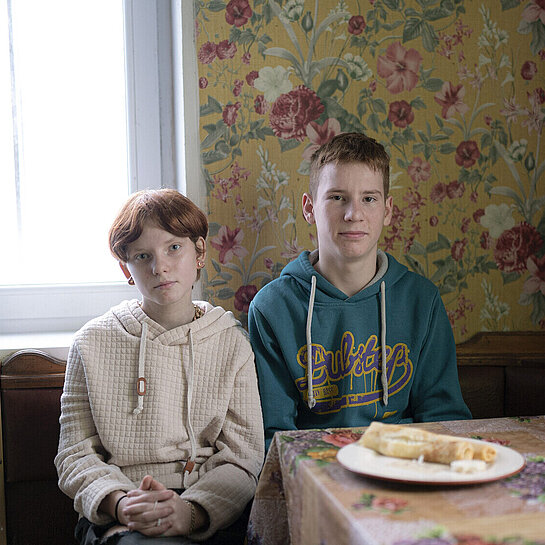 Ein Mädchen und ein Junge sitzen nebeneinander an einem Küchentisch und schauen ernst